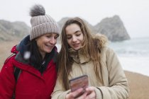 Madre e hija en ropa de abrigo usando un teléfono inteligente en la playa nevada de invierno - foto de stock