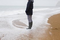Teenage boy in rubber boots standing in snowy winter ocean surf — Stock Photo