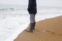 Boy in rubber boots standing at ocean surf on winter beach — Stock Photo
