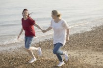 Игривая лесбийская пара, держащаяся за руки и бегущая на солнечном пляже — стоковое фото