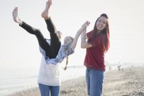 Playful lesbian couple swinging daughter on beach — Stock Photo