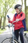 Mulher sênior ativa usando telefone inteligente na bicicleta no parque — Fotografia de Stock