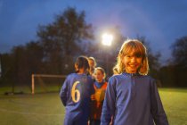 Retrato menina confiante jogando futebol com a equipe em campo à noite — Fotografia de Stock