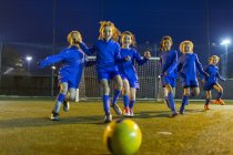 Chicas equipo de fútbol jugando, corriendo hacia la pelota en el campo por la noche - foto de stock