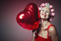 Portrait playful senior woman with hair in curlers holding heart-shape balloon — Stock Photo