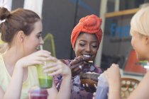 Mujeres jóvenes amigas bebiendo batidos y comiendo en la cafetería de la acera - foto de stock