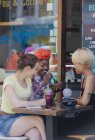 Mujeres jóvenes amigas bebiendo batidos y comiendo en la cafetería de la acera - foto de stock