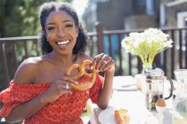 Portrait smiling, confident young woman with pretzel enjoying breakfast on sunny balcony — Stock Photo