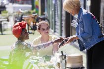 Junge Frau bezahlt Kellnerin mit Kreditkarte in sonnigem Bürgersteig-Café — Stockfoto