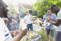 Male friends laughing and eating around barbecue grill in backyard — Stock Photo