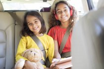Retrato irmãs felizes com ursinho montando no banco de trás do carro — Fotografia de Stock