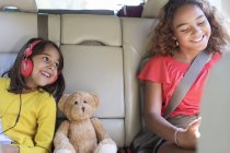 Hermanas felices y oso de peluche cabalgando en el asiento trasero del coche - foto de stock
