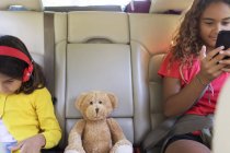 Сестры с плюшевым мишкой с помощью смартфона и цифрового планшета, катаются на заднем сиденье автомобиля — стоковое фото