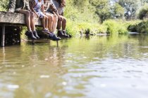 Familia colgando pies de soleado muelle junto al río - foto de stock