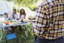 Padre sirviendo hamburguesas barbacoa a la familia en la mesa del camping - foto de stock
