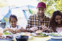Pai e filhas desfrutando de almoço hambúrguer no acampamento — Fotografia de Stock