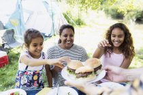 Happy mother and daughters reaching for barbecue hamburgers at campsite — Stock Photo