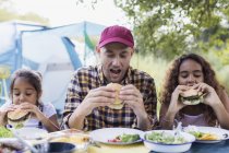 Padre e hijas comiendo hamburguesas barbacoa en el camping - foto de stock