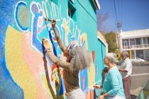 Volontari della comunità senior che dipingono vivaci murales sul soleggiato muro urbano — Foto stock