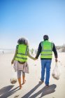 Affectionate senior couple volunteers cleaning up litter on sunny, wet sand beach — Stock Photo