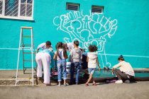 Enfants volontaires peignant une murale communautaire sur un mur ensoleillé — Photo de stock