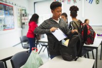 Niño de secundaria colocando cuaderno en la mochila en el aula - foto de stock