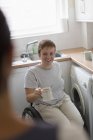 Sonriente joven en silla de ruedas bebiendo té en la cocina del apartamento - foto de stock