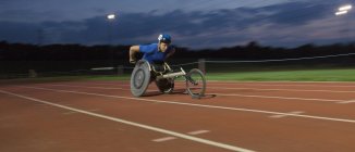 Determinato giovane atleta paraplegico maschile che accelera lungo la pista sportiva in gara su sedia a rotelle di notte — Foto stock