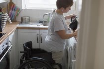 Mujer joven con silla de ruedas vertiendo té en apartamento cocina - foto de stock