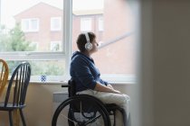 Thoughtful young woman in wheelchair listening to music with headphones at window — Stock Photo