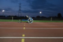 Giovane atleta paraplegica femminile che corre lungo la pista sportiva in gara su sedia a rotelle di notte — Foto stock