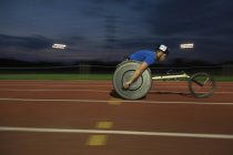 Joven macho parapléjico exceso de velocidad a lo largo de pista deportiva durante la carrera en silla de ruedas por la noche - foto de stock