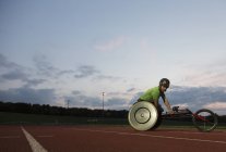 Jovem atleta paraplégico do sexo masculino treinando para corrida em cadeira de rodas em pista de esportes à noite — Fotografia de Stock