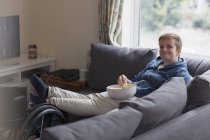 Retrato jovem confiante assistindo TV e comer pipocas no sofá com os pés para cima na cadeira de rodas — Fotografia de Stock