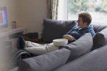 Улыбающаяся молодая женщина отдыхает на диване, смотрит телевизор и ест попкорн с ногами на инвалидной коляске — стоковое фото
