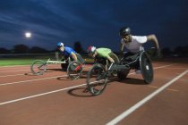 Atletas parapléjicos corriendo a lo largo de pista deportiva en silla de ruedas en la noche - foto de stock
