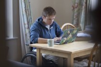 Сосредоточенная молодая женщина в инвалидной коляске с ноутбуком за обеденным столом — стоковое фото