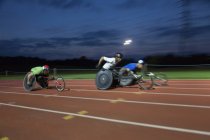 Atletas parapléjicos corriendo a lo largo de pista deportiva en carrera en silla de ruedas por la noche - foto de stock