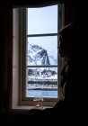 Pensive woman looking out window at snowy mountain, Reine, Lofoten Islands, Norway — Stock Photo