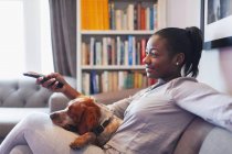 Jovem mulher e cão relaxante, assistindo TV no sofá da sala de estar — Fotografia de Stock