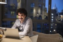 Hombre sonriente retrato usando portátil en apartamento urbano por la noche - foto de stock