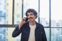 Улыбающийся мужчина разговаривает по смартфону у окна городской квартиры — стоковое фото