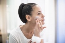 Woman applying moisturizer to face in bathroom mirror — Stock Photo