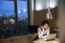 Hombre usando laptop y bebiendo vino blanco en apartamento urbano por la noche - foto de stock
