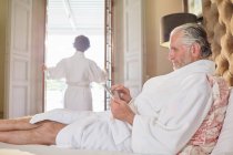 Homme mûr en peignoir de spa en utilisant une tablette numérique sur le lit de l'hôtel — Photo de stock