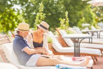 Couple relaxant, livre de lecture et utilisation du téléphone intelligent sur chaise longue au bord de la piscine de l'hôtel — Photo de stock