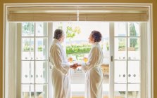 Mature couple in spa bathrobes drinking mimosas at hotel patio door — Stock Photo