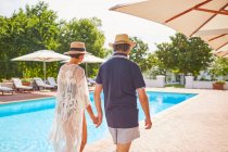 Зріла пара тримає руки, прогулюючись вздовж сонячного курортного басейну — стокове фото