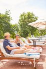 Mature couple relaxing, reading on lounge chairs at sunny resort poolside — Stock Photo