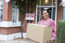 Portrait smiling, confident woman moving into new house, holding cardboard box outside house — Stock Photo